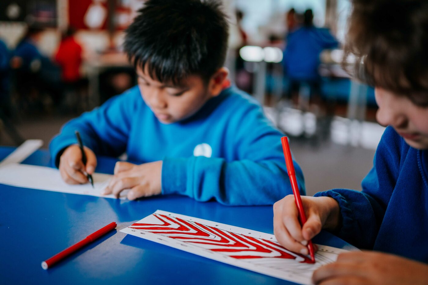 Two children colouring in patterns on some paper at a desk.
