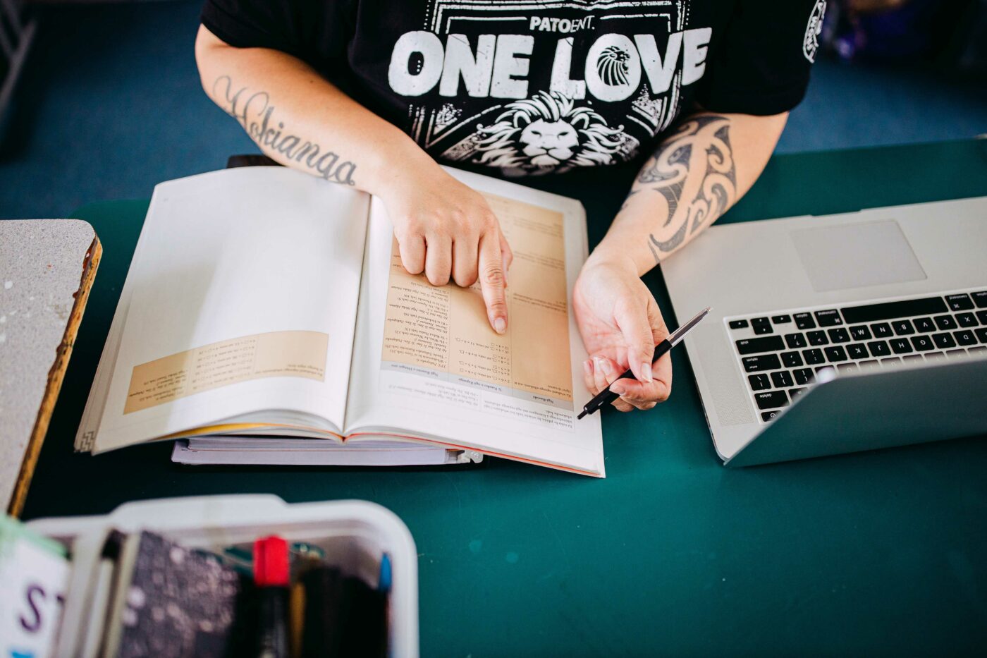 Hands pointing at a line in a text book, next to a laptop on a desk.