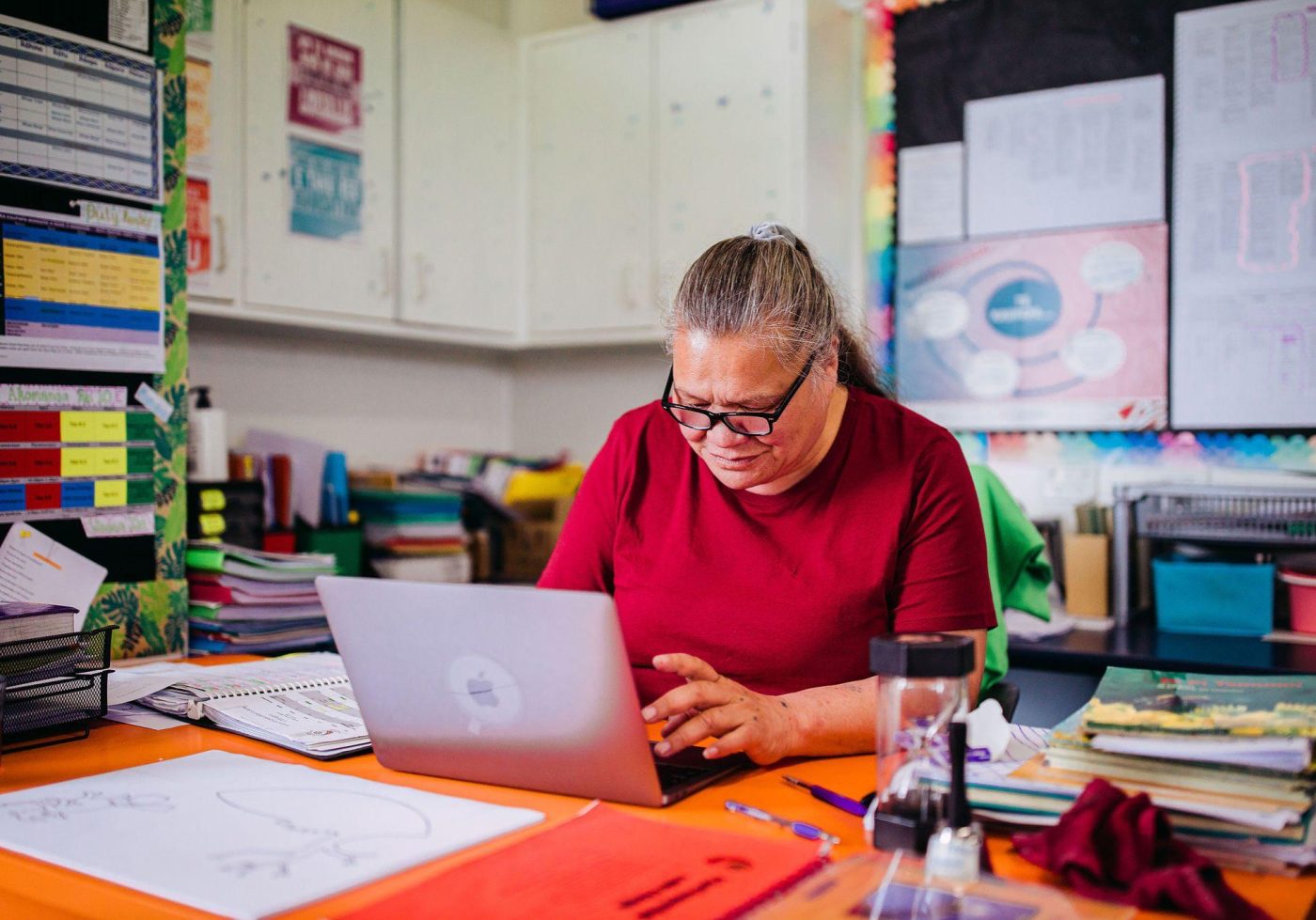 A woman with grey hair and a red shirt sits at a desk working on a laptop.