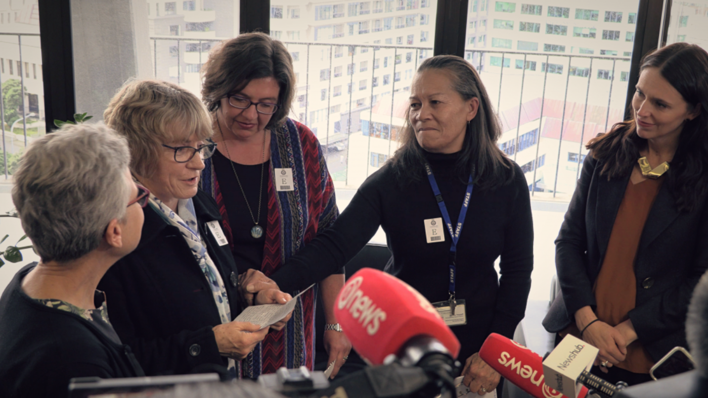 Four women stand talking to media at the beehive with Prime Minister Jacinda Ardern looks on. Cameras and media microphones are visible in the bottom of the shot.