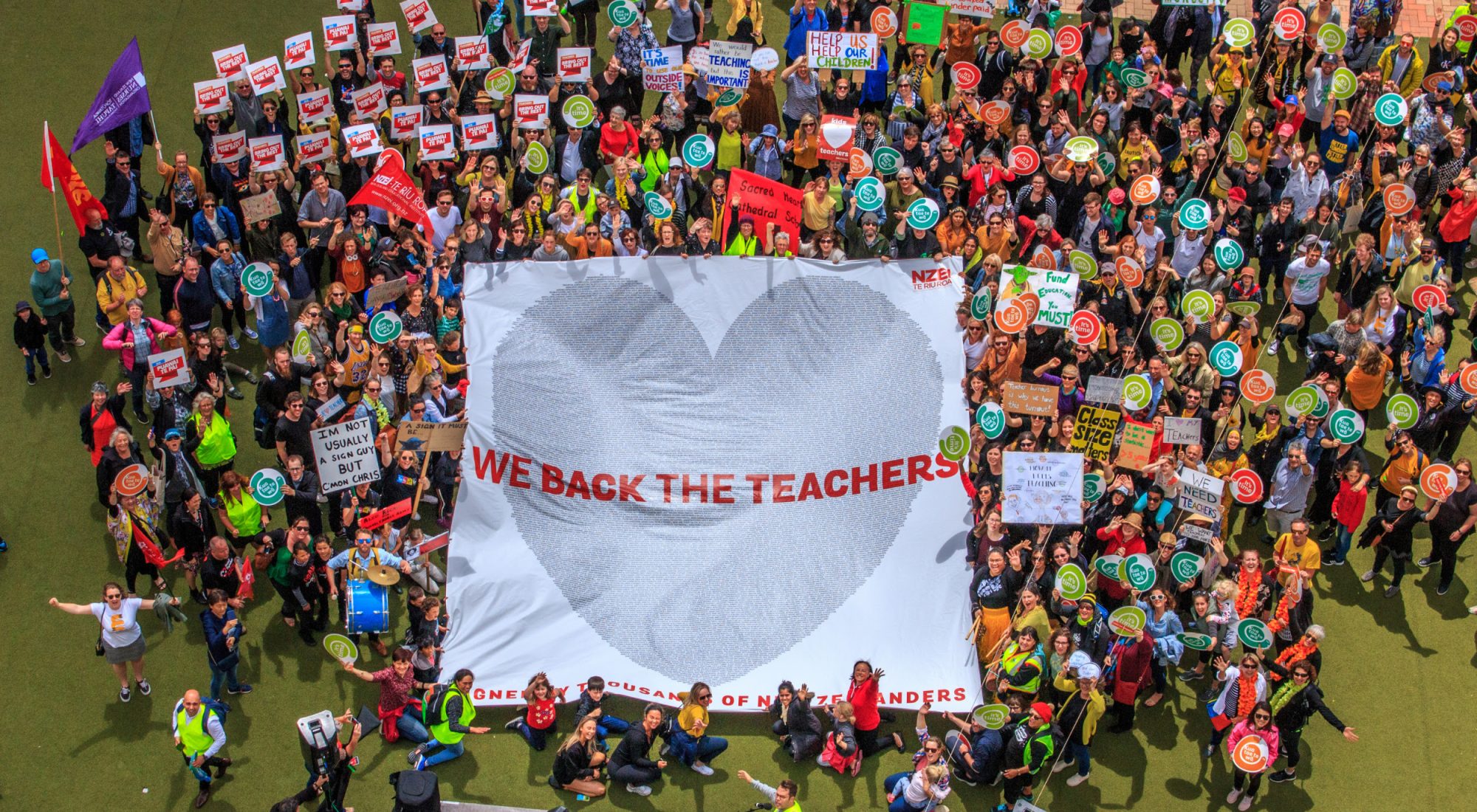 Arial shot of hundreds of members gathered in Civic Square holding a giant banner. Banner reads: "We back the teachers."